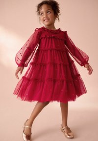 Giovane ragazza che sorride indossando un vestito rosso a balze con lunghe maniche trasparenti ricamate e scarpe Mary Jane dorate, in piedi su uno sfondo beige.