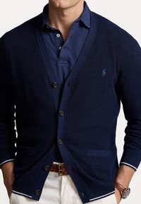 Marinblå cardigan med en strukturerad väv, fem knappar, två fickor och vita detaljer vid ärmarna, bärs över en marinblå polo. 
