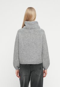 En blond kvinne står med ryggen synlig, iført en lys grå tykkstrikket genser med høy hals og mørke grå jeans med lommer på lapp.