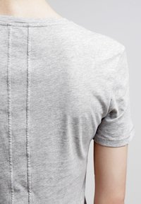 Detailní záběr na horní část zad osoby, která nosí světlou šedou tričko s viditelným vertikálním švem uprostřed.