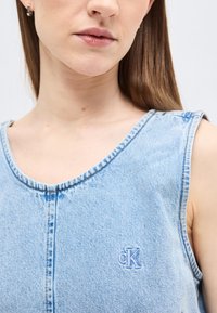 Κοντινό πλάνο μιας γυναίκας που φοράει αμάνικο ανοιχτό μπλε τζιν τοπ με μικρό κεντημένο λογότυπο «cK» στο στήθος.
