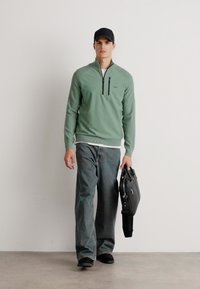 Grüner Reißverschluss-Pullover mit hohem Kragen, graue Jeans, schwarze Schuhe und eine schwarze Tasche. Das Modell steht vor einem neutralen Hintergrund.