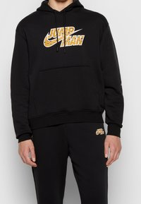 Sort pullover-hoodie med kængurulomme, der har et gult Nike-logo med dyreprint på fronten. Blødt materiale i bomuldsblanding.