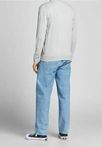 Person som bär en ljusgrå långärmad tröja, löst sittande ljusblå jeans och marinblå sneakers, står vänd bort mot en enfärgad grå bakgrund.