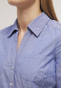 Blå knappar skjorta med en texturerad tyg, spetskrage och två bröstfickor. Designen är skräddarsydd med subtila sömdetaljer.