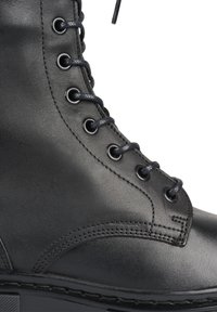 Bota de combate de cuero negro con cordones redondos, ojales de metal y costuras en contraste. Suela acolchada y acabado suave.
