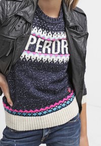 Person, der einen bunten Strickpullover mit dem Schriftzug "PERD" trägt, kombiniert mit einer schwarzen Lederjacke und blauen Jeans, mit der hand auf der Hüfte.