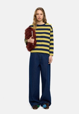 Kobieta w żółto-granatowej paskowanej bluzce z długim rękawem, szerokich ciemnych jeansach, trzyma bordową pluszową kopertówkę, stoi na jasnoszarym tle.