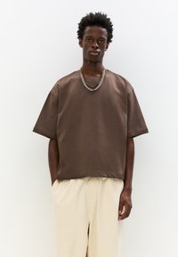 Übergroßes braunes Baumwoll-T-Shirt mit V-Ausschnitt und kurzen Ärmeln, kombiniert mit hellbeigen, locker sitzenden Hosen und einer geschichteten Kettenhalskette.