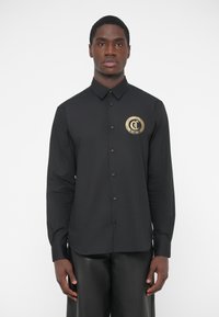 Camisa negra de manga larga con botones, con un logo circular dorado en el pecho izquierdo, textura lisa, cuello clásico y botones negros.