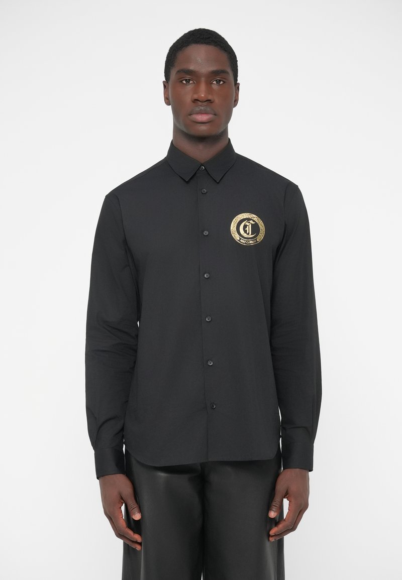 Camisa negra de manga larga con botones, con un logo circular dorado en el pecho izquierdo, textura lisa, cuello clásico y botones negros.