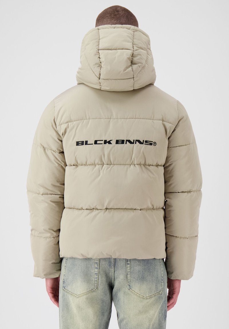 Pufferjack in lichtbeige met een gewatteerd ontwerp, voorzien van een grote capuchon en een zwart "BLCK BNNS" logo op de achterkant.