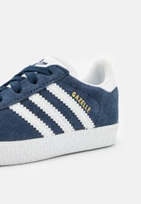 Navy blue suede sneaker with white leather stripes and gold "Gazelle" text. White rubber sole features a textured pattern for grip.