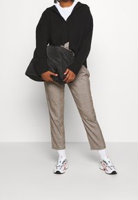 Svart stickad zip-up hoodie, grå rutiga byxor, vita strumpor och flerfärgade sneakers. Väskan är svart och puffy, hålls i ena handen.