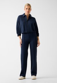 Giacca blu navy con cerniera, colletto e maniche lunghe, abbinata a pantaloni aderenti coordinati. Tessuto morbido e liscio con dettagli di design minimi.