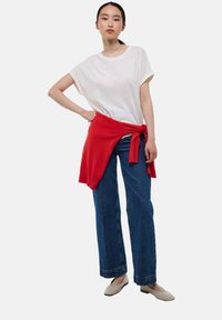 T-shirt blanc à manches courtes associé à un jean bleu taille haute, un pull rouge noué à la taille et des ballerines beiges. Tenue simple et décontractée.