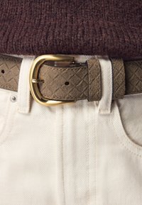 Textured brown belt with a gold buckle, worn over white denim. The belt features a diamond pattern and is fastened through a loop.