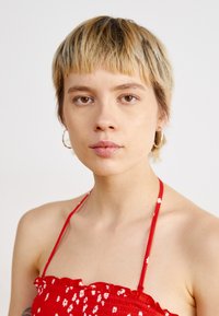 Červený vyhrnutý crop top s kvetinovým vzorom, tenkými pásikmi a volánkovým výstrihom. Modelka s krátkymi vlasmi a kruhovými náušnicami. Jednoduché pozadie.