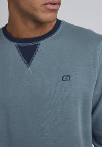 Lysblå sweatshirt med en ribbet marineblå krave, der har kontrastsyer og et lille broderet logo på brystet.