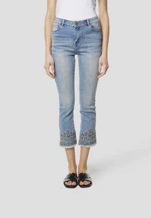 Femme portant un jean court bleu clair avec des ourlets brodés et des sandales noires, debout devant un fond uni.