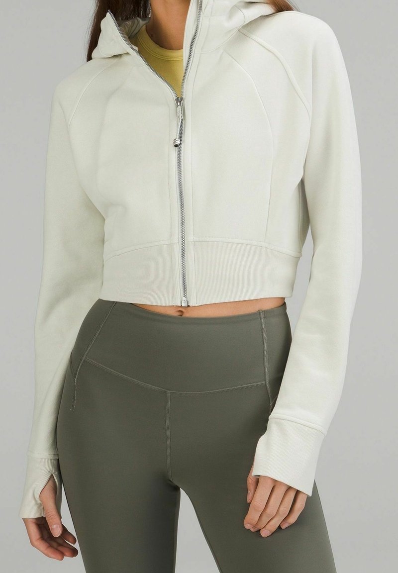 Femme portant un sweat à capuche zippé court écru avec des trous pour les pouces et des leggings taille haute vert foncé sur un fond neutre.