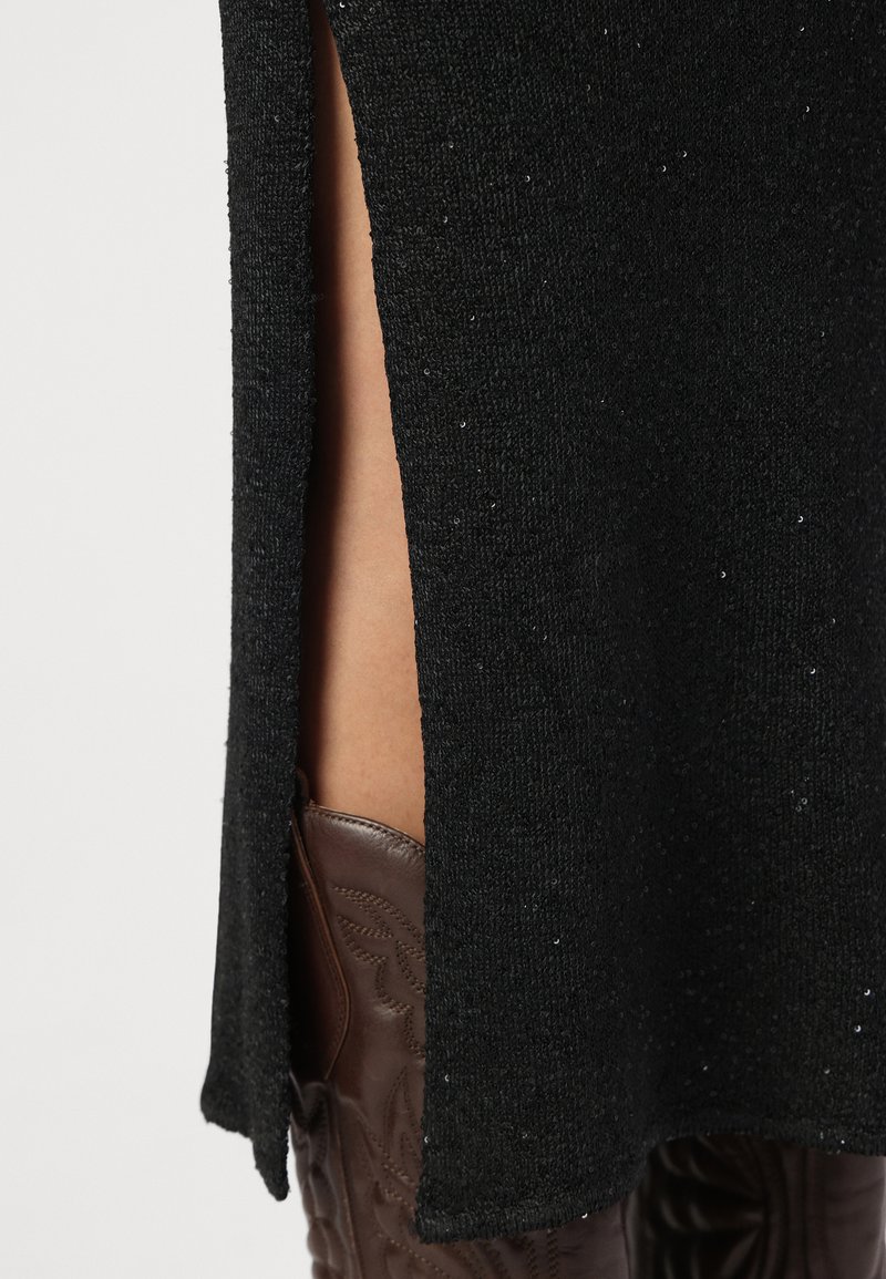 Schwarzes gestricktes Kleid mit seitlichem Schlitz, aus strukturiertem Stoff mit dezentem Glanz, kombiniert mit braunen, dekorativen Stiefeln im Westernstil.