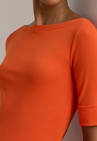 Oranžs ribots tops ar laivu veida kaklu, pieguļoša dizaina un elkoņu garuma piedurknēm. Gluda tekstūra ar nelielu spīdumu.