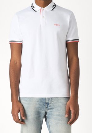 Man draagt een wit poloshirt met zwart-roze gestreepte bies aan de kraag en mouwen, met het "BOSS"-logo op de linkerborst, gecombineerd met lichte jeans.