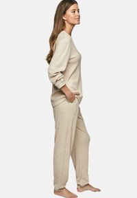 Beige geribbelde lounge-outfit bestaande uit een langarmtop en wijdvallende broek, beide met zakken en een zachte textuur, gedragen op blote voeten.