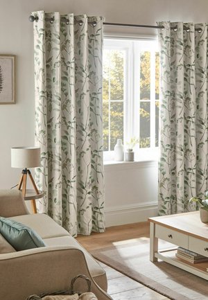 Wohnzimmer mit floral gemusterten Vorhängen, beigem Sofa mit blauem Kissen, hölzernem Couchtisch, Lampe auf einem Dreibeinständer und Fenster mit Blick ins Grüne.