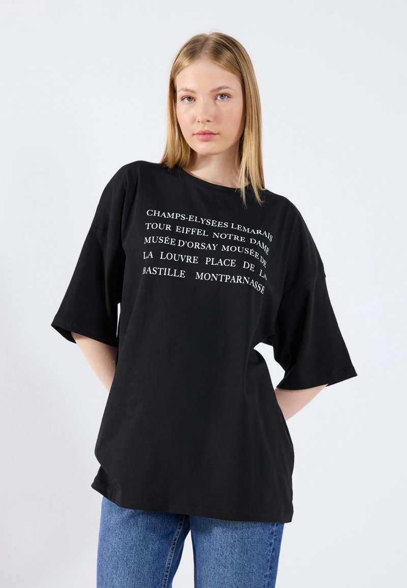Camiseta negra extragrande de algodón con texto blanco que enumera monumentos de París. Mangas cortas y un corte relajado.