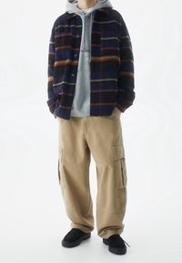 Persona che indossa una giacca a righe navy con bottoni sopra un hoodie grigio, pantaloni cargo beige e scarpe nere, in piedi con le mani in tasca.