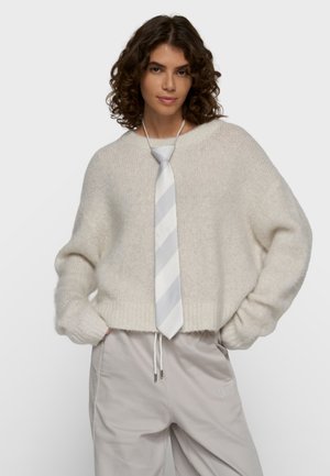 Kvinde med krøllet hår iført en løs cremefarvet sweater, lyst gråstribet slips og lysebeige bukser med hænderne i lommerne.