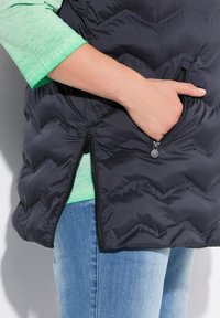 Schwarze Steppweste mit Chevron-Muster, mit seitlichen Reißverschlusstaschen, darunter ein grünes T-Shirt und kombiniert mit blauen Jeans.