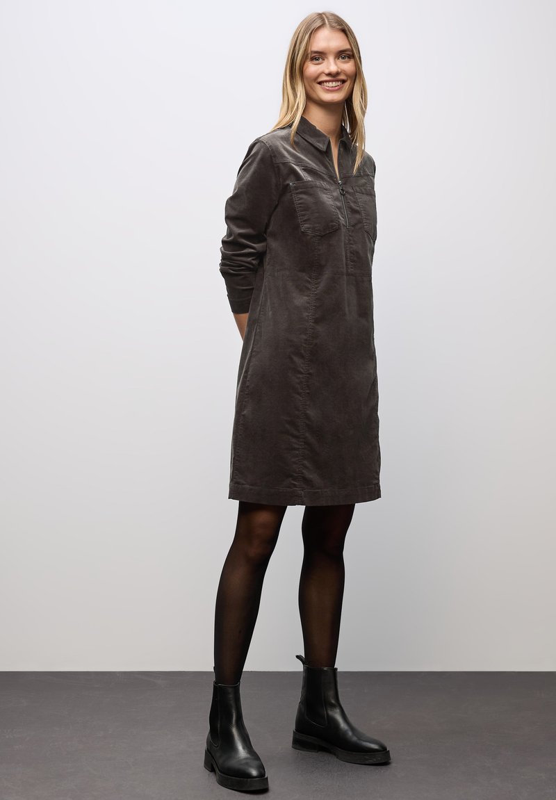 Donkergrijze corduroy jurk met kraag, lange mouwen, voorzak en een zoom boven de knie. Gemaakt in combinatie met enkellaarsjes.