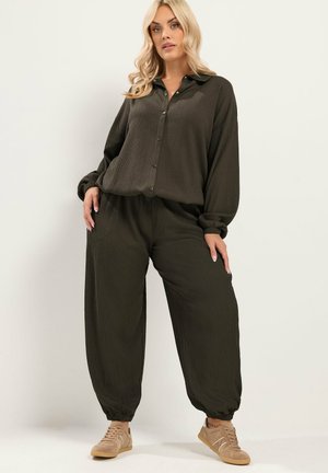 Blond vrouw met een losse donkerolijfgroene blouse met knopen en bijpassende wijde broek met beige sneakers, staand tegen een effen lichte achtergrond.