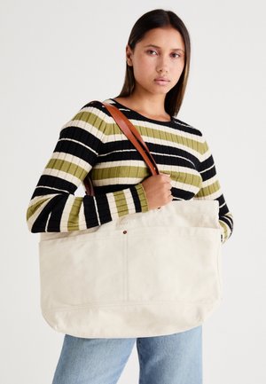 Grande borsa in tela beige con manici in pelle marrone, caratterizzata da un design minimal e dettagli di cucitura visibili.