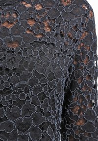 Svart spetstyg med tät blommönster som ligger ovanpå genomskinlig mesh, som visar intrikata stygn och delikat genomskinlighet.