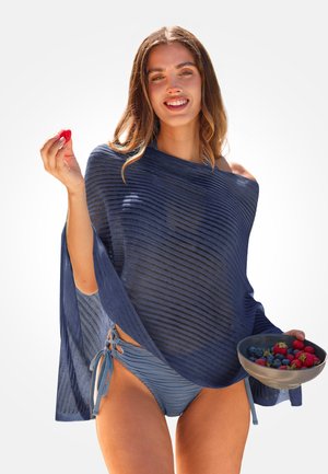 Mujer joven con traje de baño azul y chal sonriendo, sosteniendo un tazón de frutas del bosque mixtas y una baya en la mano.