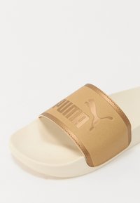 Liukkaat sandaalit, joissa on leveä kultainen hihna, jossa on kohopainettu "PUMA"-logo ja hyppivä puuma -kuvio, asetettuna karhealle beigeelle pohjalle.