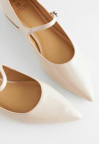 Ballerines en cuir verni beige avec un bout pointu, une bride au niveau de la cheville et une texture lisse. L'intérieur présente une semelle intérieure marron contrastante.
