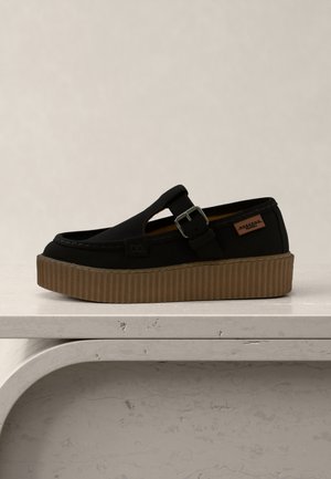 Sort slip-on sko med brun platformsål, sidespænde og lille lædermærke med mærke, vist på en tekstureret hvid overflade.