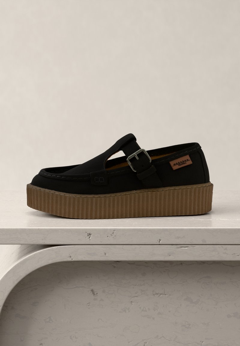 Chaussure noire à enfiler avec une semelle plateforme marron, une lanière latérale à boucle et une petite étiquette de marque en cuir, présentée sur une surface blanche texturée.