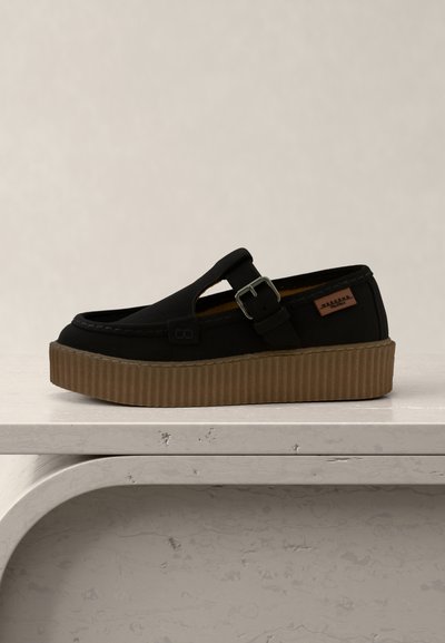 Chaussure noire à enfiler avec une semelle plateforme marron, une lanière latérale à boucle et une petite étiquette de marque en cuir, présentée sur une surface blanche texturée.