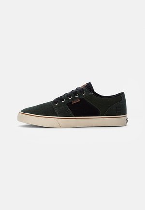 Black and green suede sneaker with a low profile, featuring a beige sole, flat laces, and a small brown logo detail on the side.
