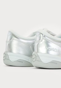 Zapatillas deportivas plateadas que presentan un material suave y reflectante, con un panel lateral texturizado, detalles en gris suave y un diseño de suela curvada.
