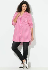 Lichtroze-wit gestreept lang overhemd met knoopsluiting aan de voorkant en borstzak, gecombineerd met zwarte skinny jeans en zwart-witte sneakers.