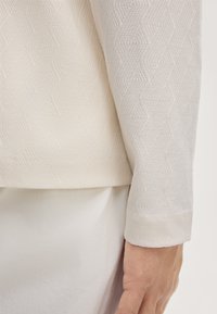 Pullover aus einem strukturierten, hellbeigen Stoff mit einem dezenten geometrischen Muster. Der Ärmelbund hat eine glatte Oberfläche. Der Hintergrund ist neutral.