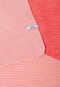 Textielstuk in roze en koraal, met een geribbeld textuur en een rand met rafelingen. Een etiket met de naam "CECIL" is zichtbaar.