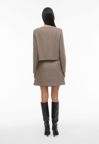 Smeđa karirana skraćena jakna uparena s odgovarajućom suknjom, upotpunjena crnim kožnim čizmama do koljena. Glatka tekstura, strukturirani dizajn.